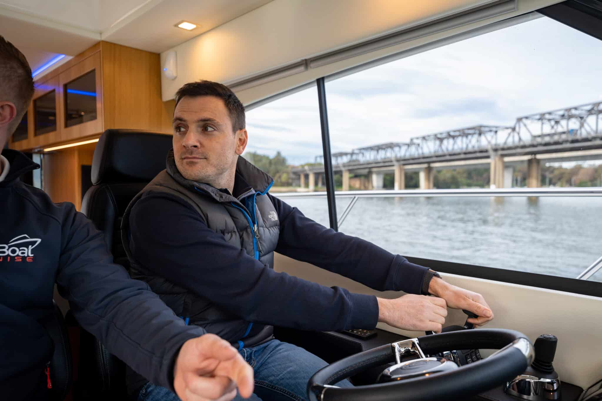 A person steers a boat on a calm river, with a bridge visible through the window. Another person is partially visible to the left.