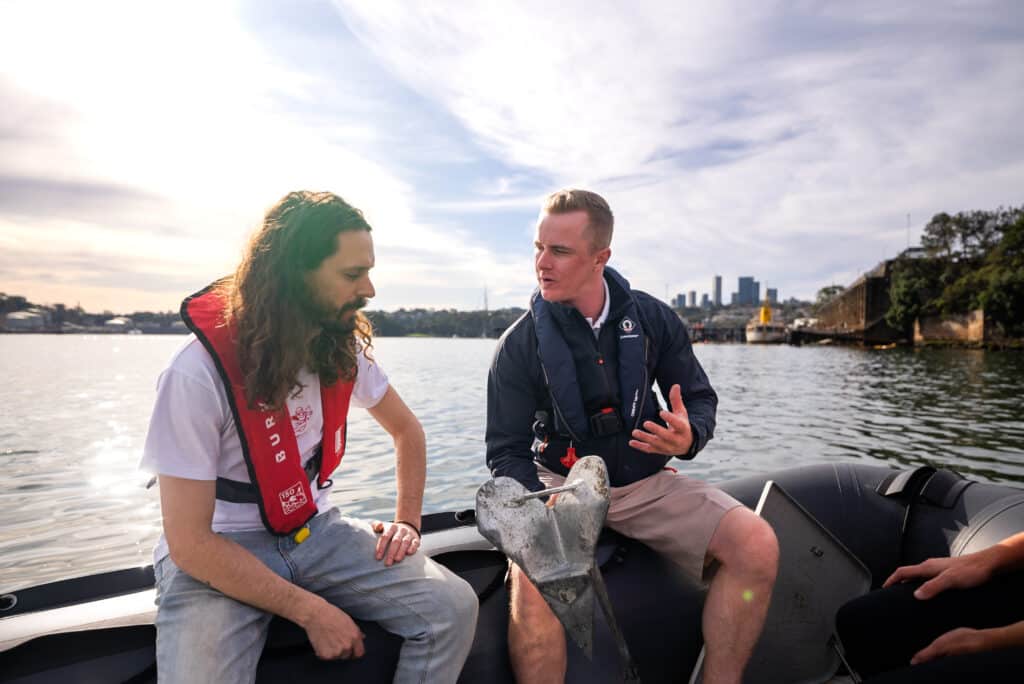 Two people sit in a boat discussing something. Both wear life vests. The person on the right gestures with their hand. Water and a city skyline are visible in the background.