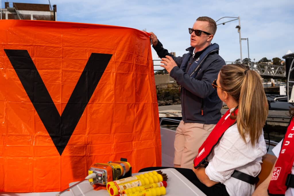 A man shows a large orange flag with a black "V" to a woman on a boat, with safety equipment visible in the foreground.