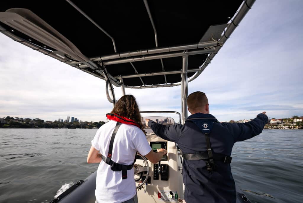 Two people on a boat; one steering, wearing a life vest, and the other pointing at something. Water and a cityscape are visible in the background.