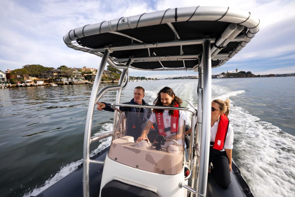 Three people on a motorboat in a calm body of water; one person at the steering wheel, the others seated. They all wear life jackets. Coastal houses and trees are visible in the background.
