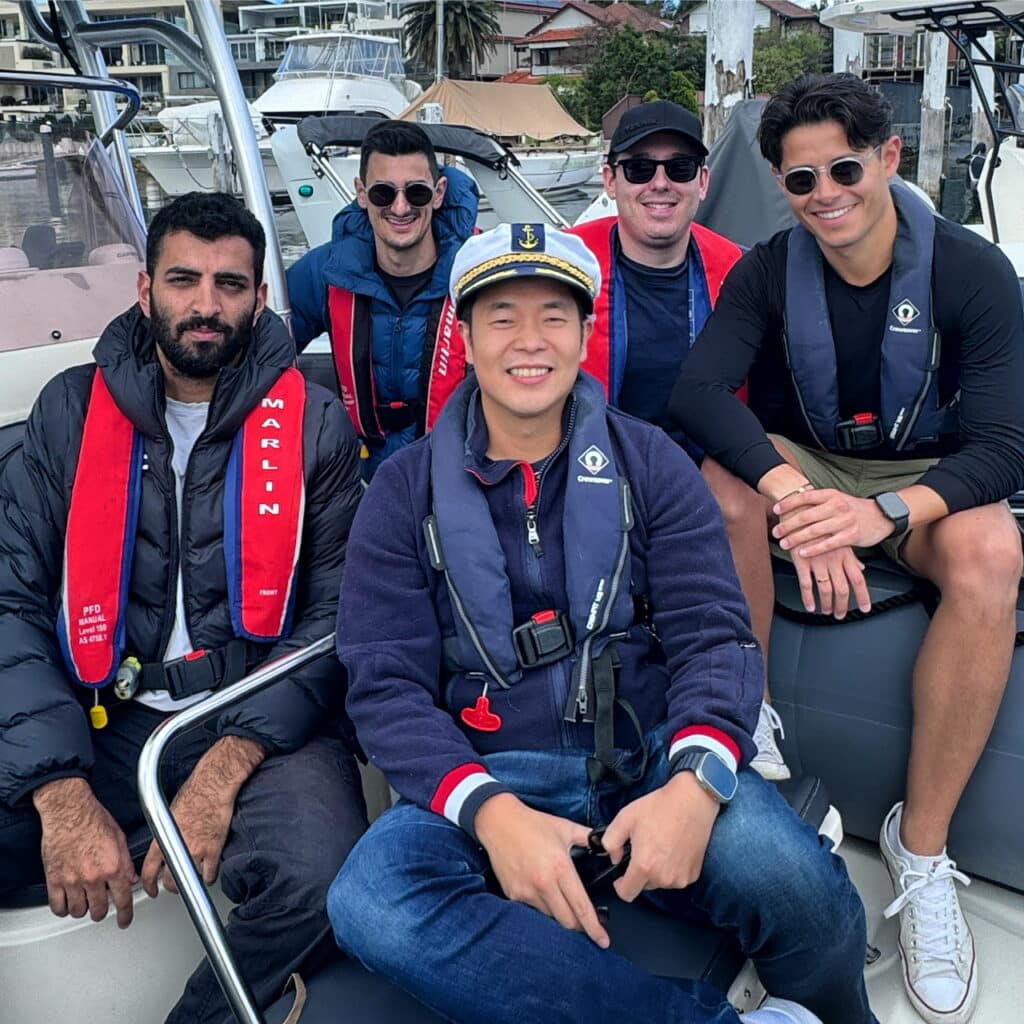 Five men wearing life jackets sit together on a boat, docked at a marina with buildings and palm trees visible in the background.
