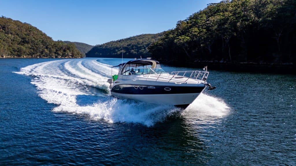 A white motor yacht speeds across a lake or river, creating a wake behind it, with forested hills in the background under a clear blue sky.