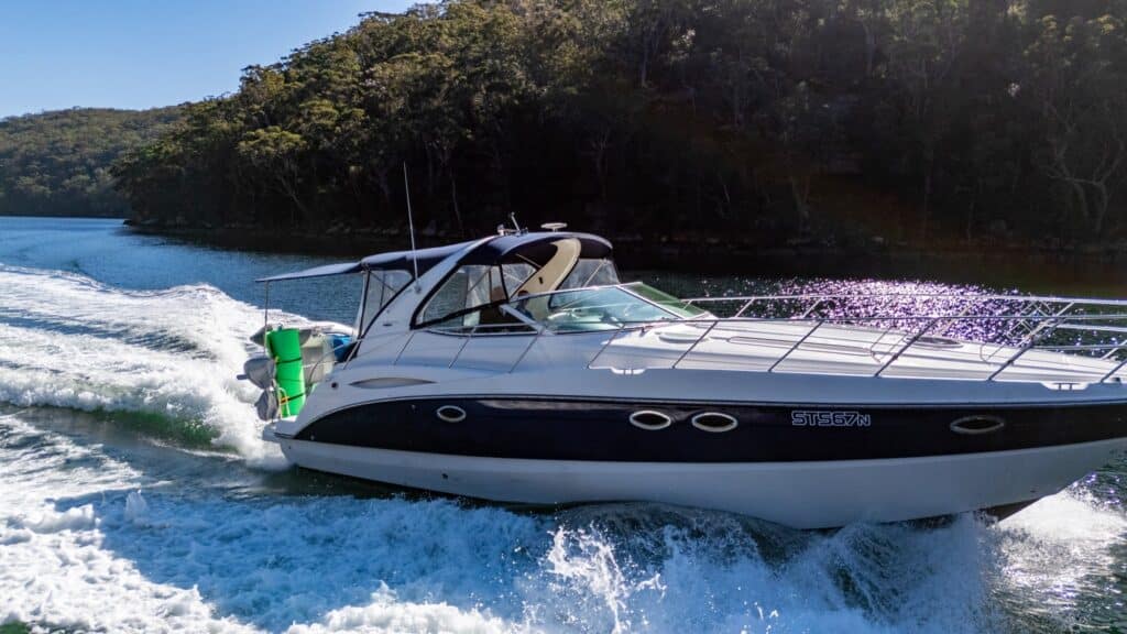 A white and black motorboat speeds through the water near a forested shoreline, creating waves behind it on a sunny day.