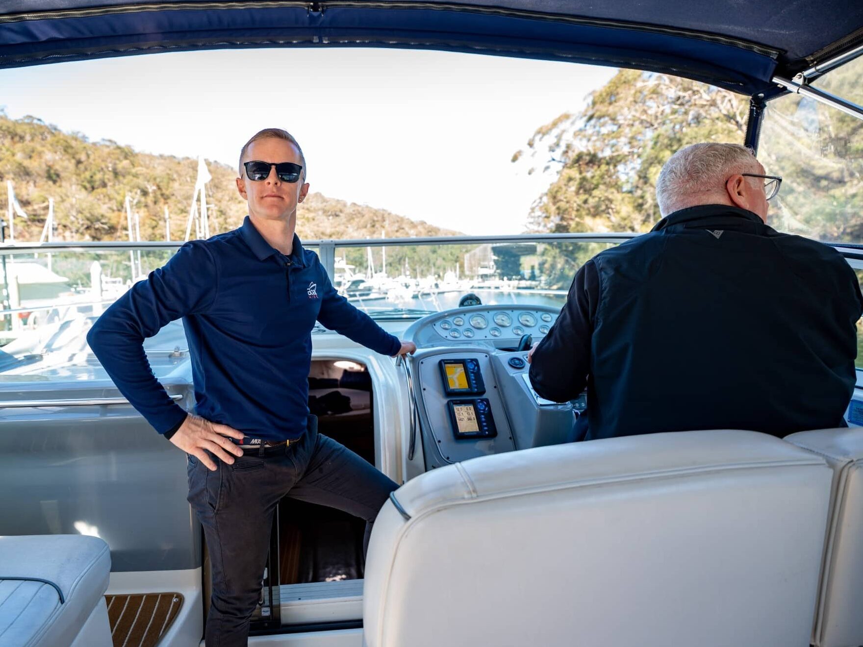 Two men are on a boat; one is standing by the controls wearing sunglasses, while the other is seated at the helm. Trees and other boats are visible in the background.