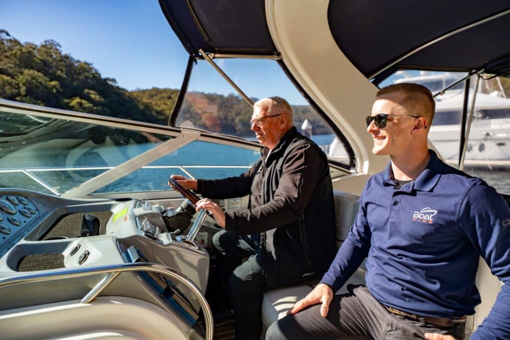 Two men are seated in the cockpit of a boat; one is steering while the other sits beside him, both facing forward on a sunny day near the water.