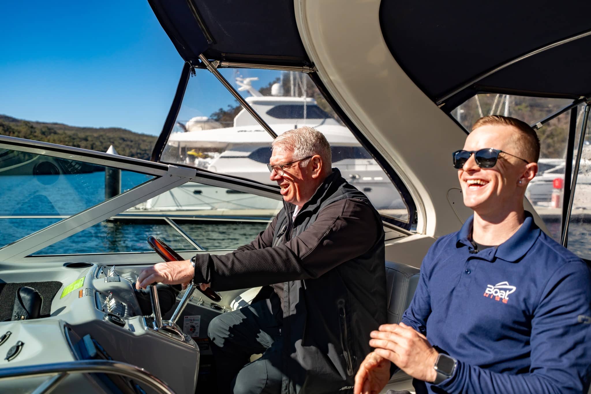 Two men on a boat, one steering and the other smiling, with yachts and water visible in the background on a sunny day.