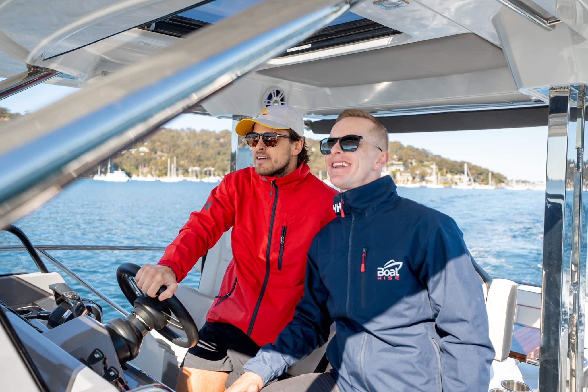 Two men wearing jackets and sunglasses are at the helm of a boat on a sunny day, with water and boats visible in the background.