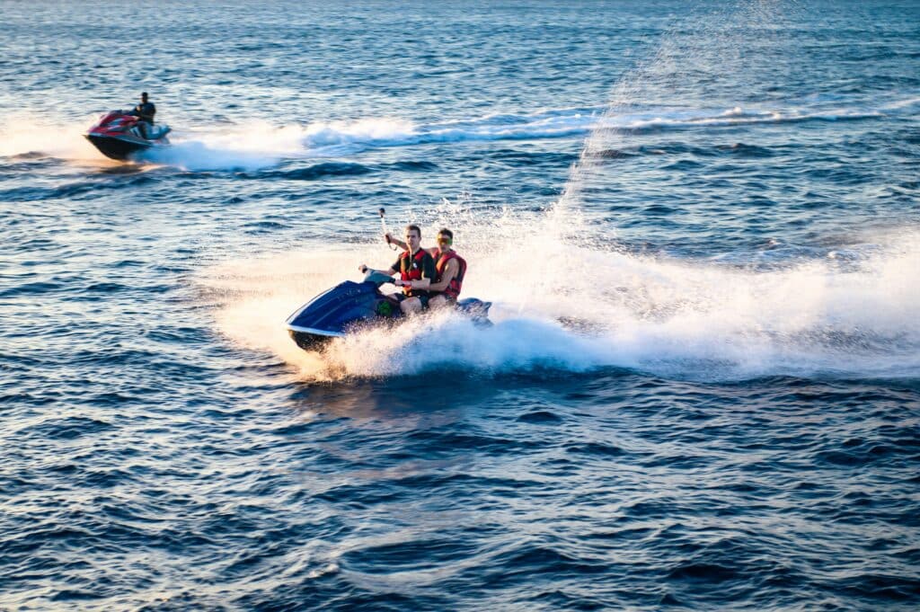 Two people ride a jet ski on the ocean, creating a spray of water, while another person rides a separate jet ski in the background.