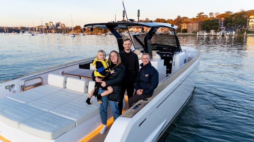 Four people, including a child in a yellow life jacket, are standing and sitting on a white motorboat in calm waters near a marina with several docked boats and buildings in the background.
